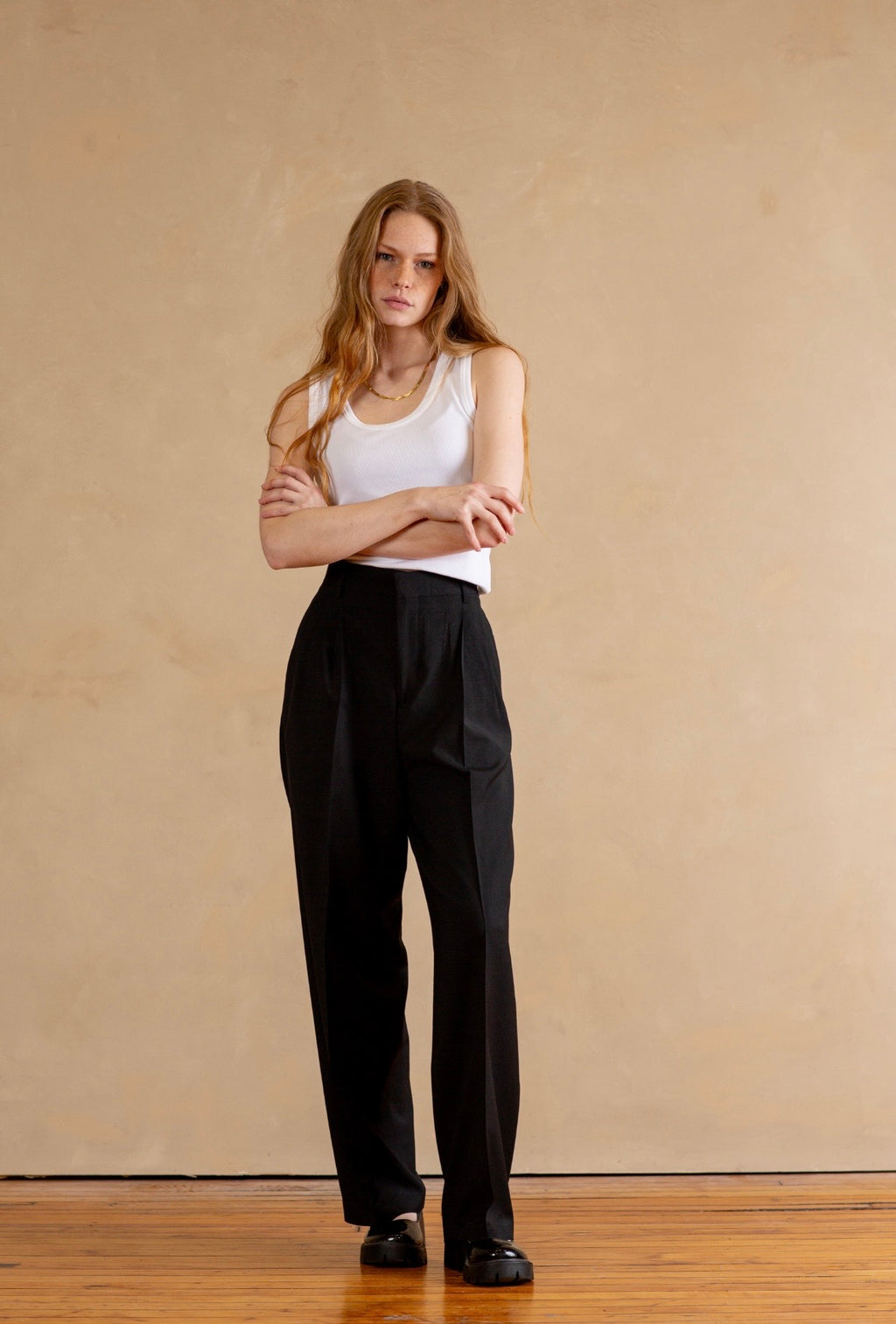 Woman wearing a white tank top and black pants standing against a beige wall.