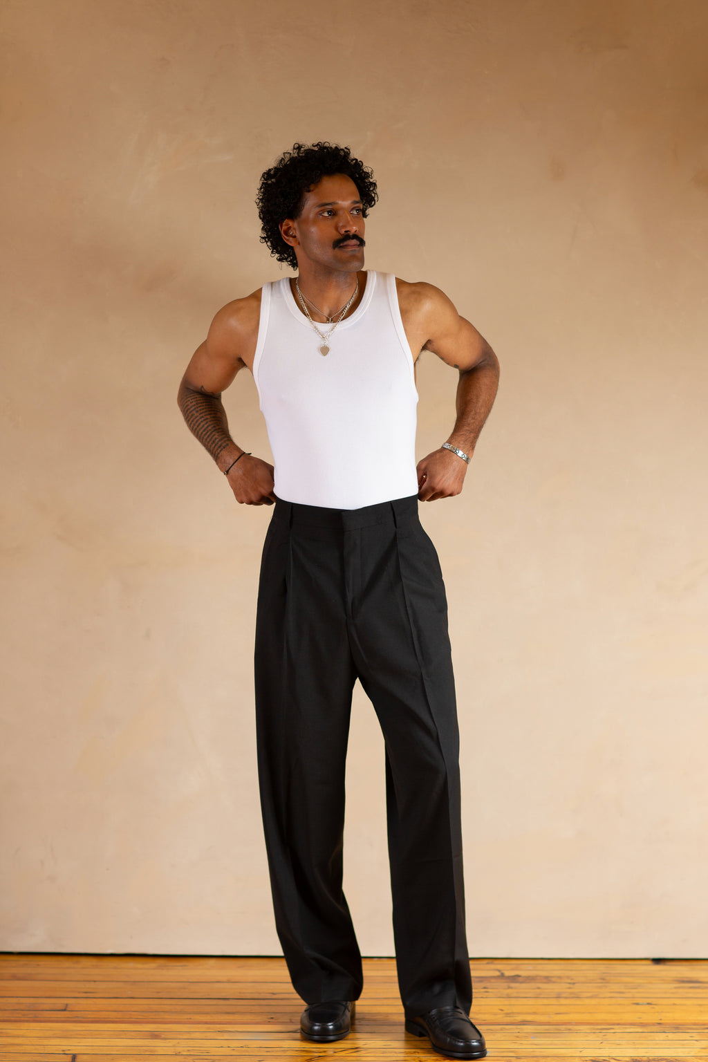 Man wearing a white tank top and black pants against a beige wall.