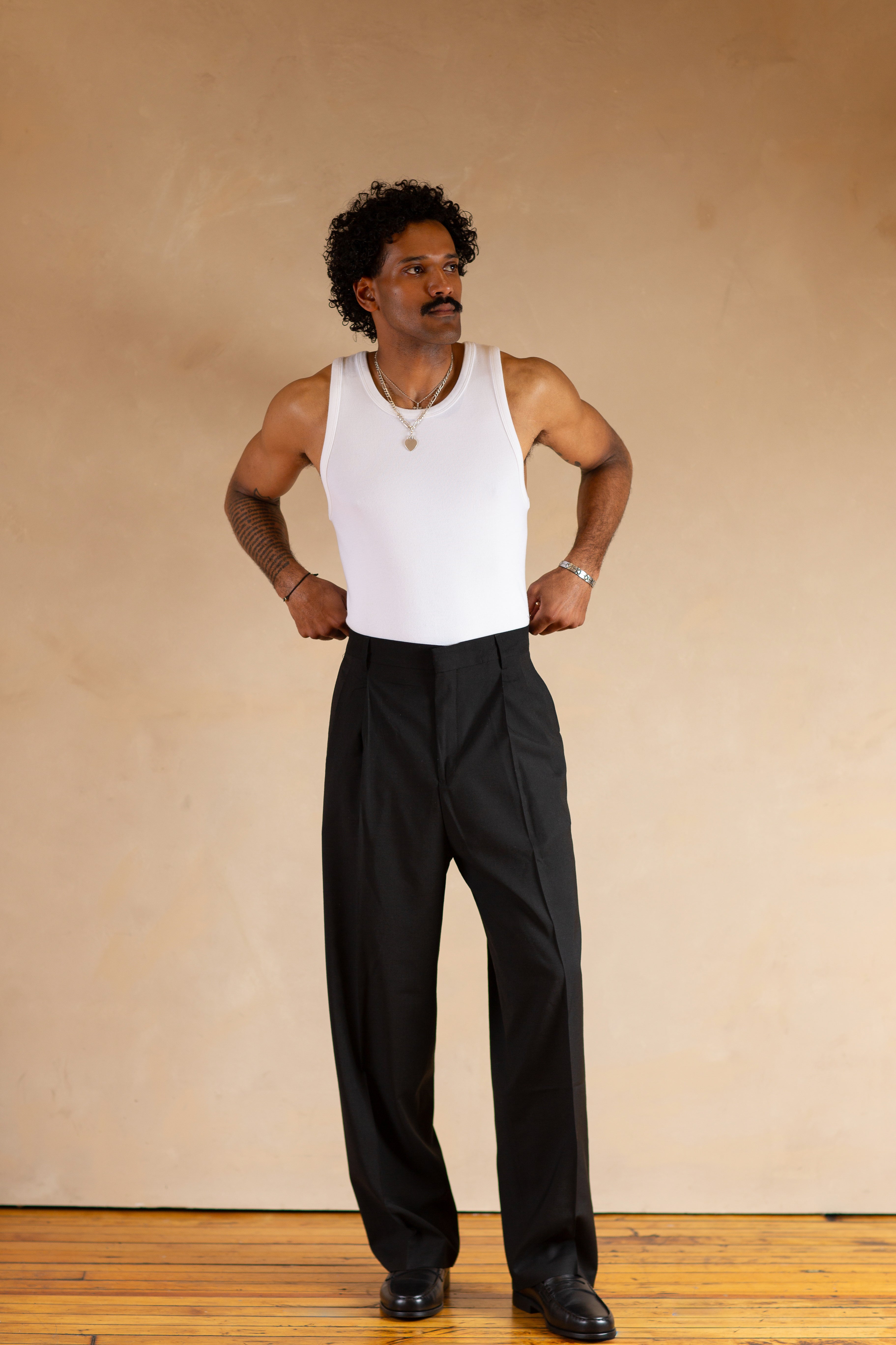 Man wearing a white tank top and black pants against a beige wall.