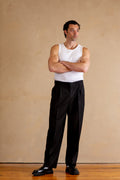 Man wearing a white tank top and black pants standing against a beige wall.