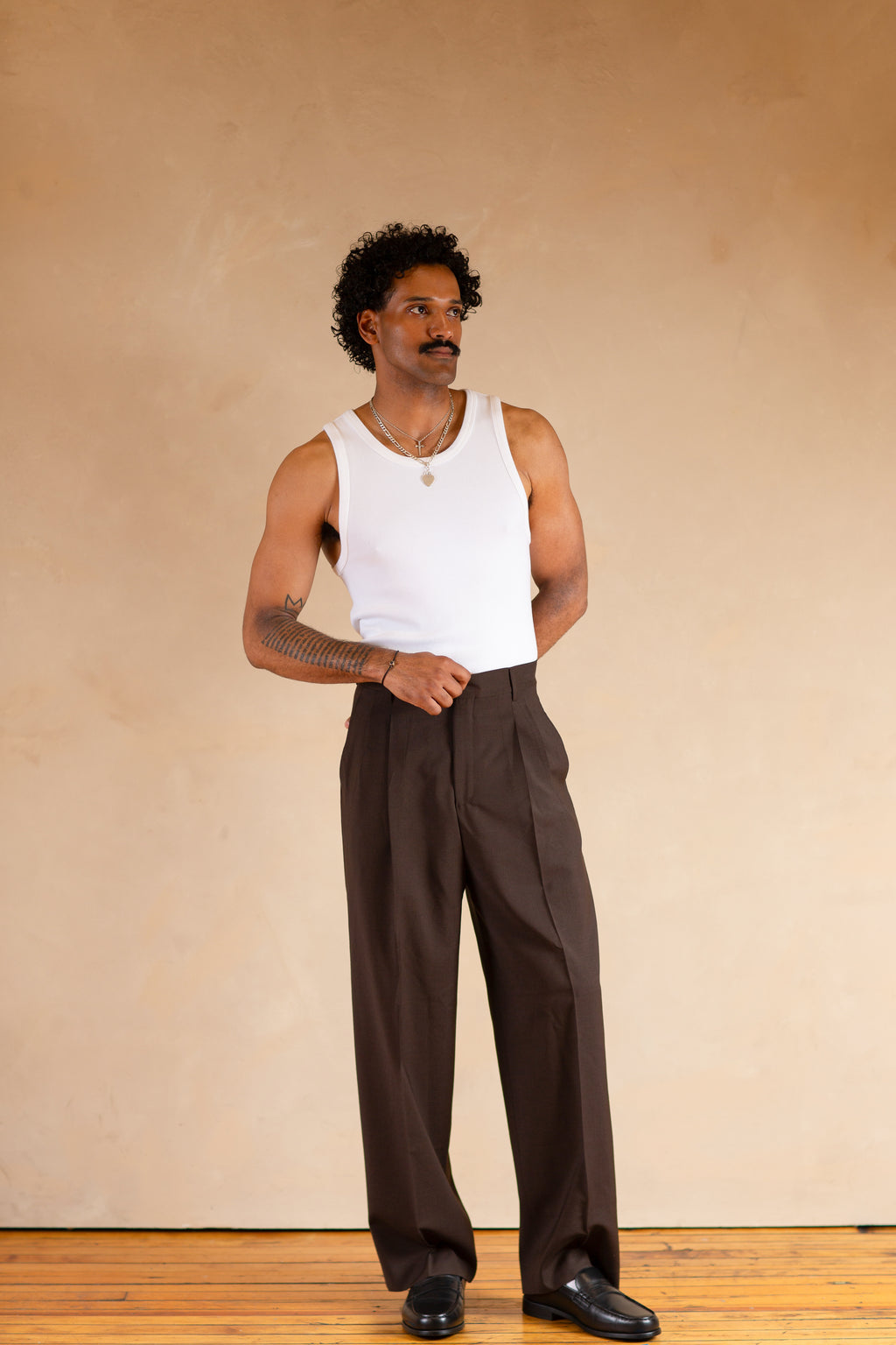 Man wearing a white tank top and brown pants standing against a beige wall.