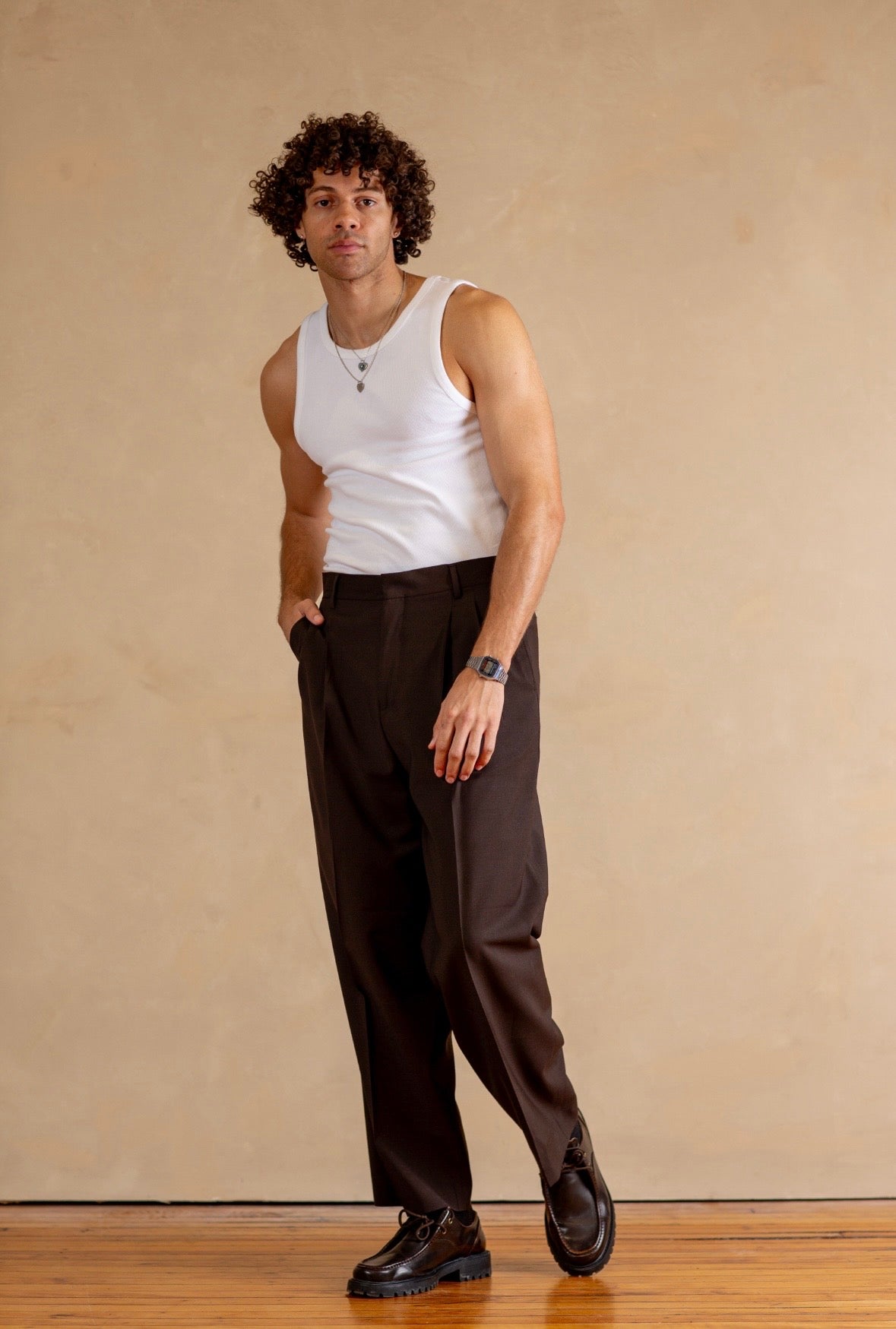 Person wearing a white tank top and brown pants against a beige wall.