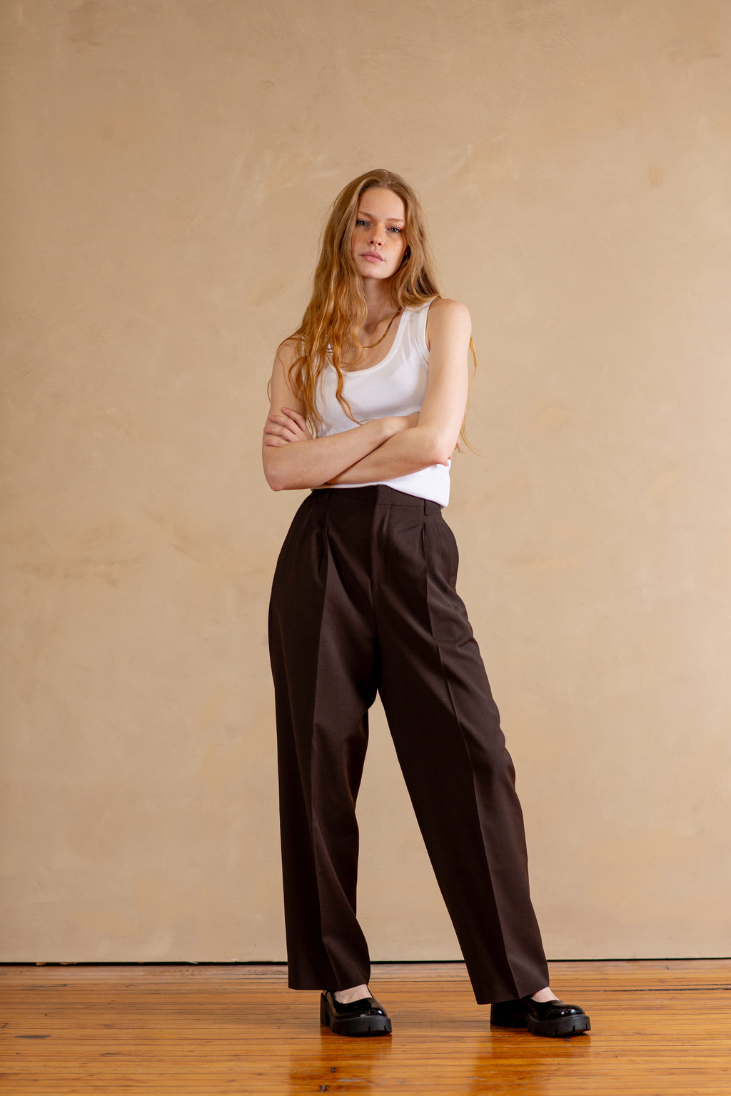 Woman wearing a white tank top and brown pants standing against a beige wall.