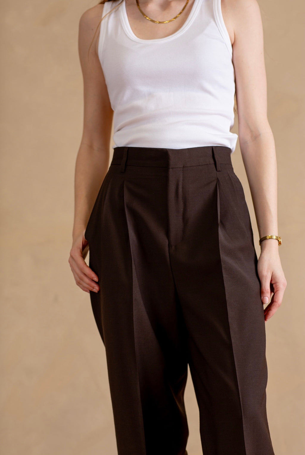 Person wearing a white tank top and brown pleated pants on a beige background