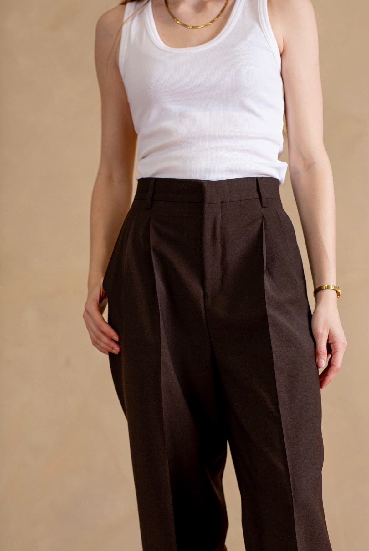 Person wearing a white tank top and brown pleated pants on a beige background