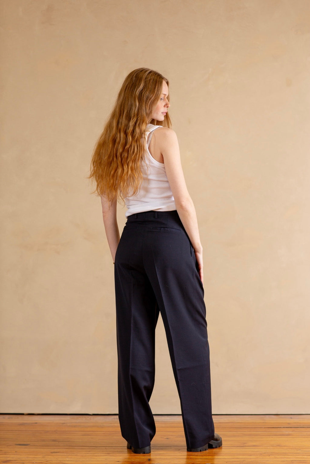 Woman wearing a white tank top and navy pants against a beige wall.