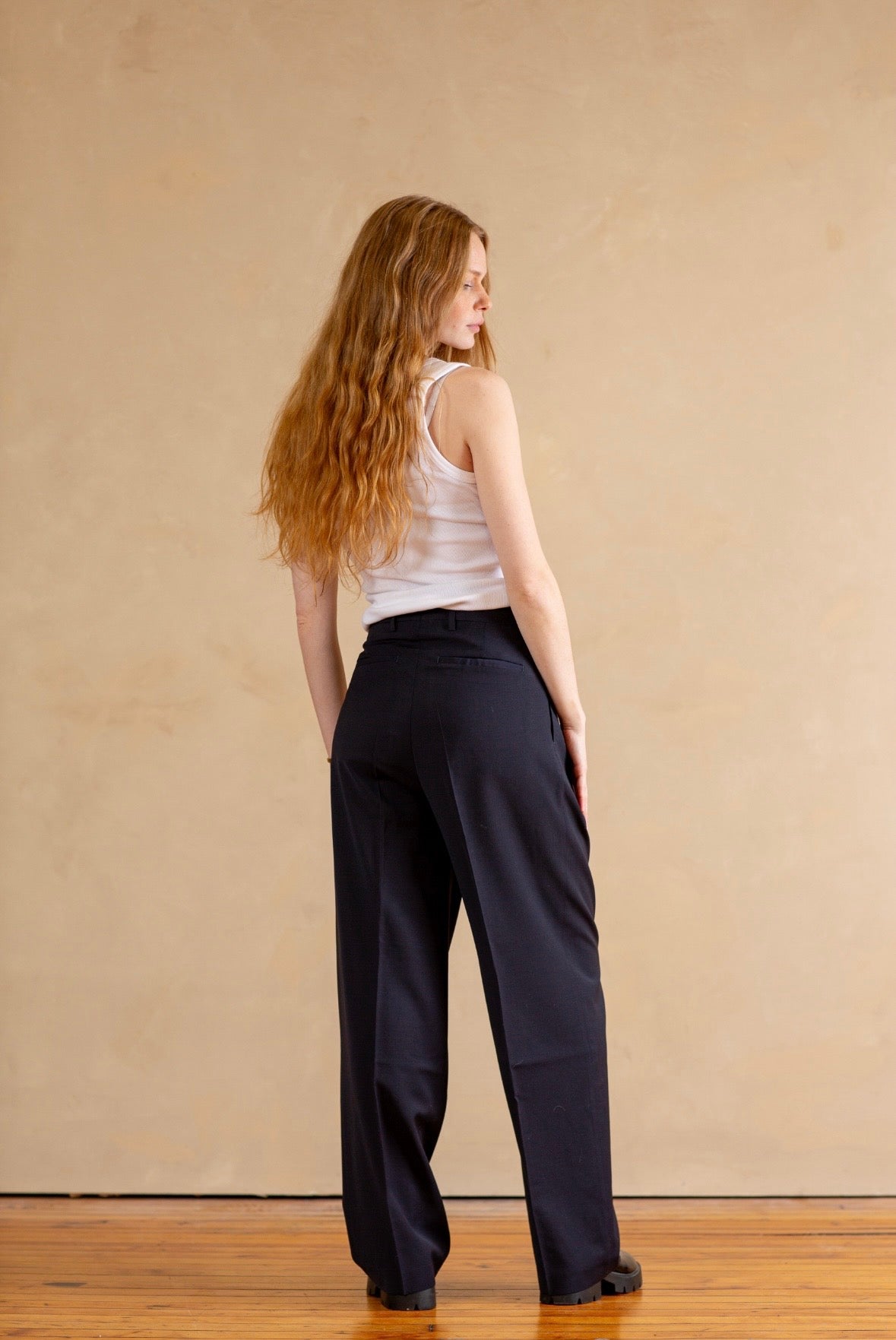 Woman wearing a white tank top and navy pants against a beige wall.