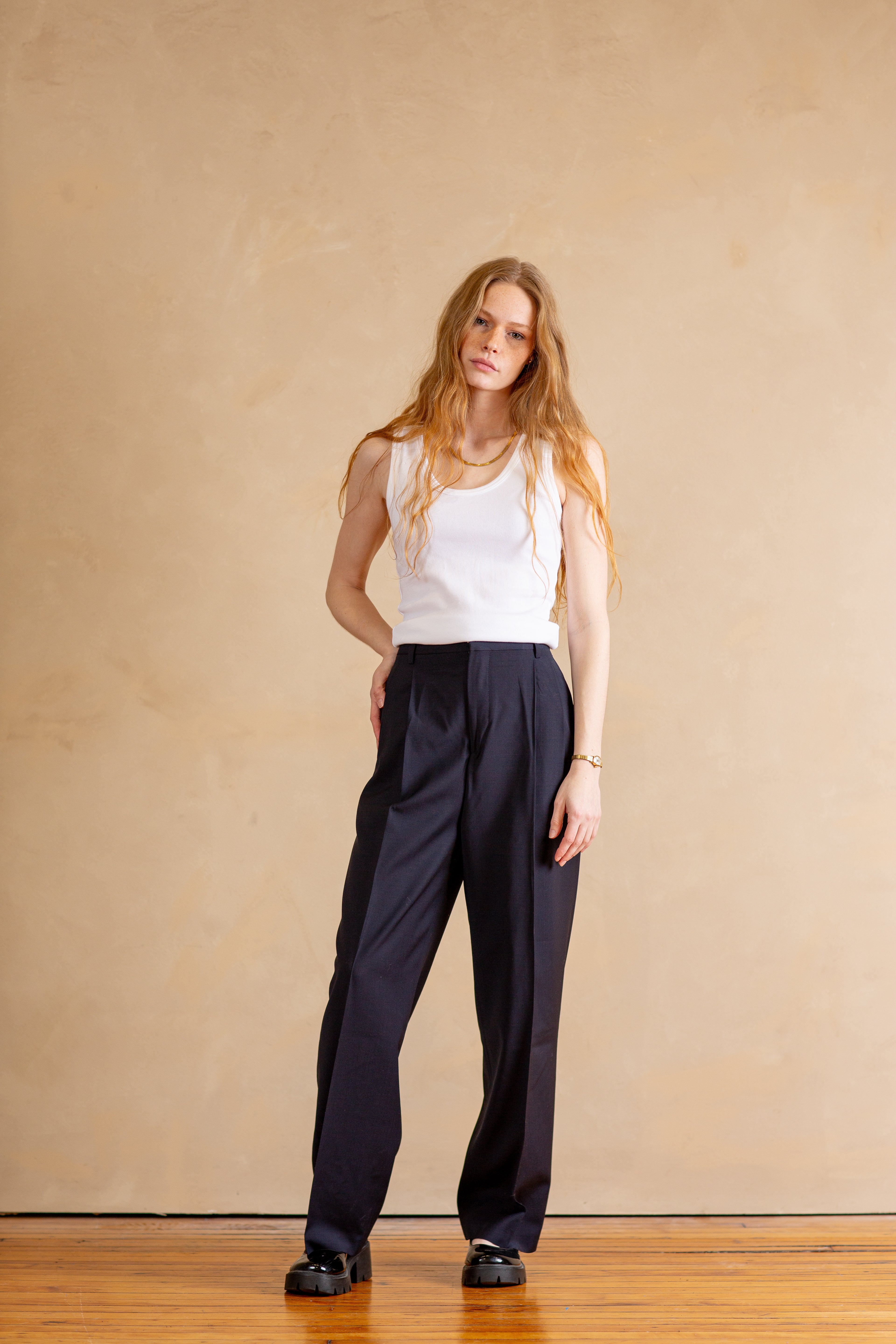 Woman wearing a white sleeveless top and navy pants against a beige wall.