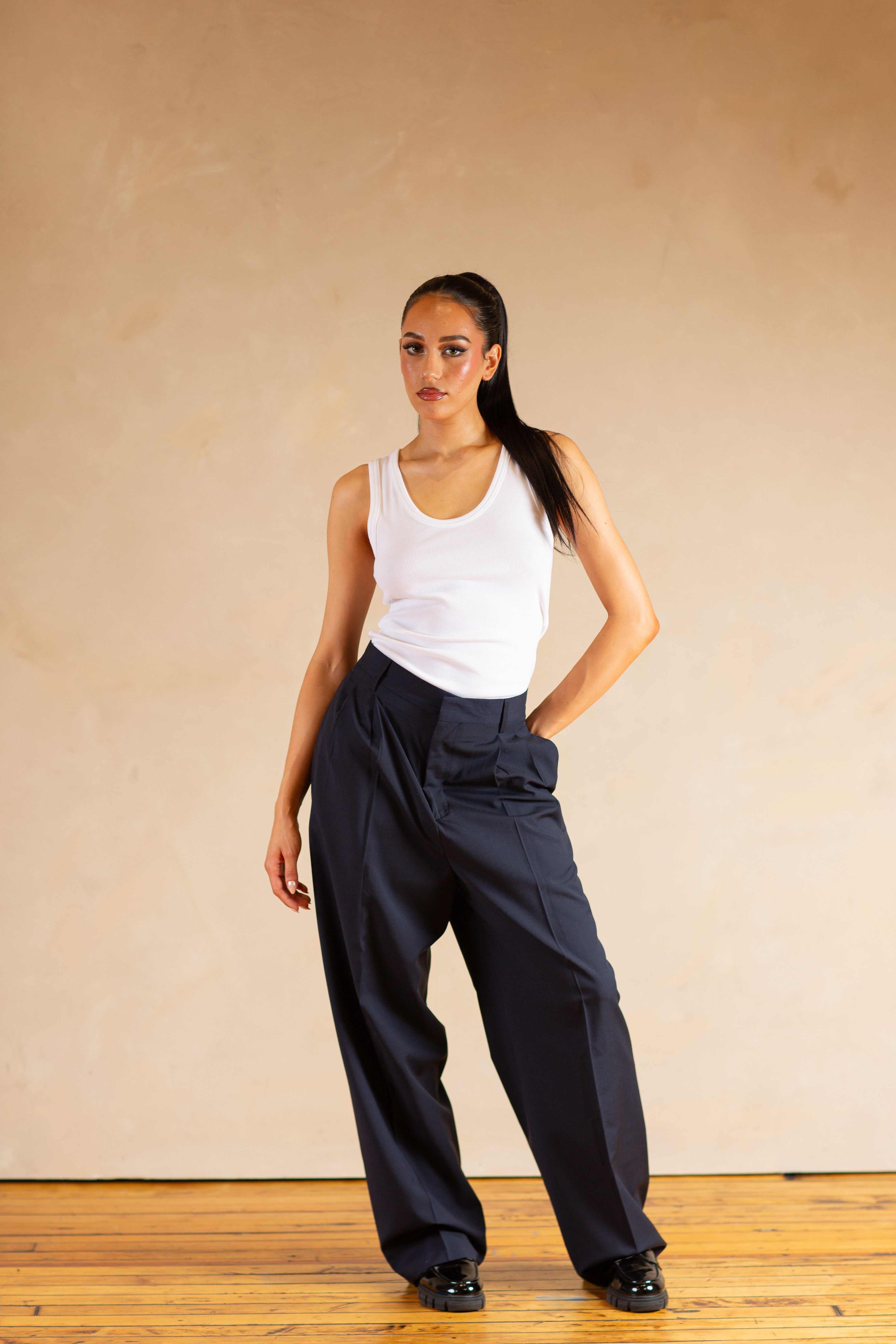 Woman wearing a white tank top and Navy pants against a beige wall.