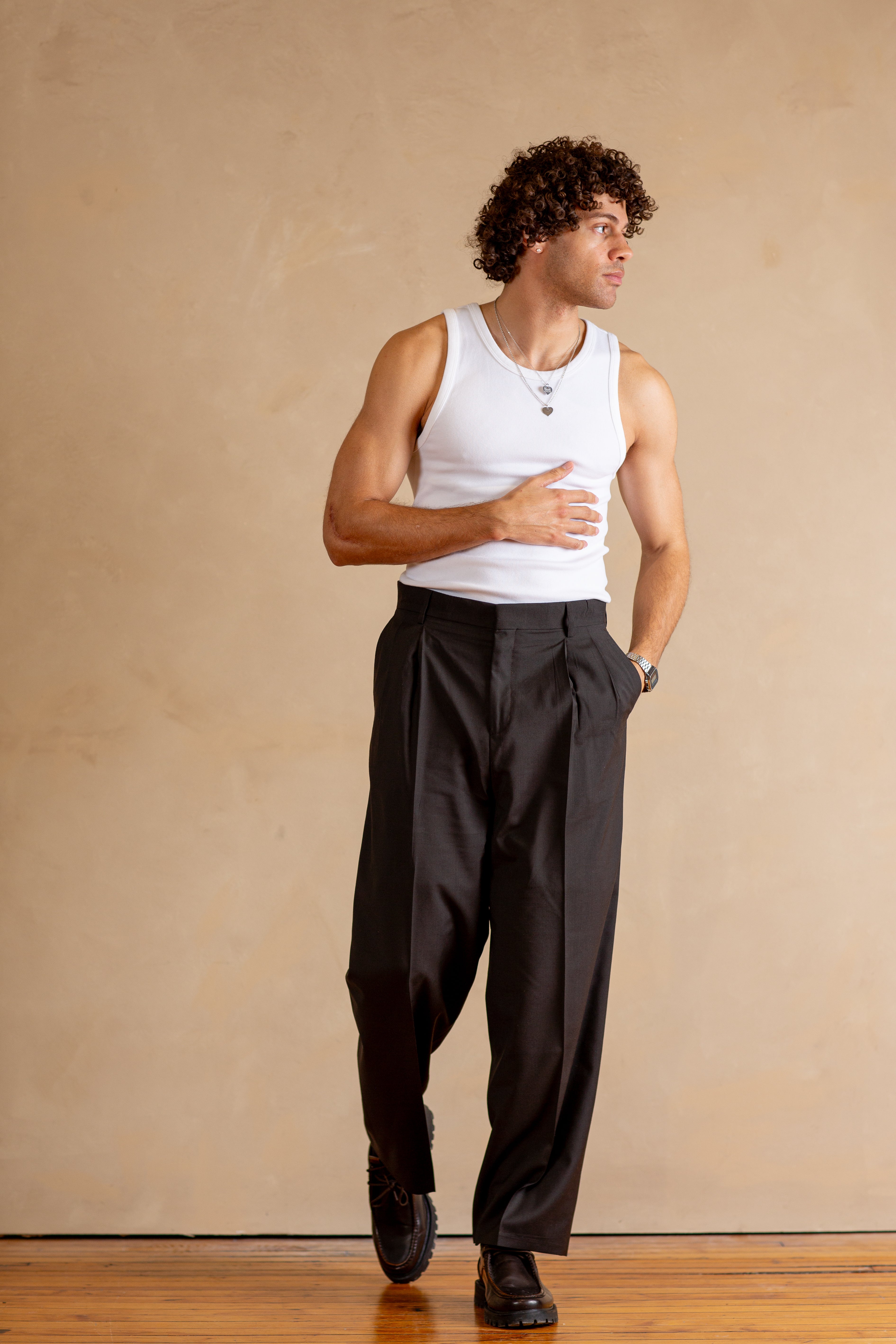 Man wearing a white tank top and Stone pants against a beige wall.
