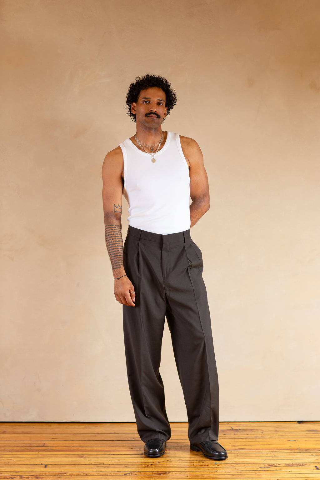 Man wearing a white tank top and Stone pants standing against a beige wall.