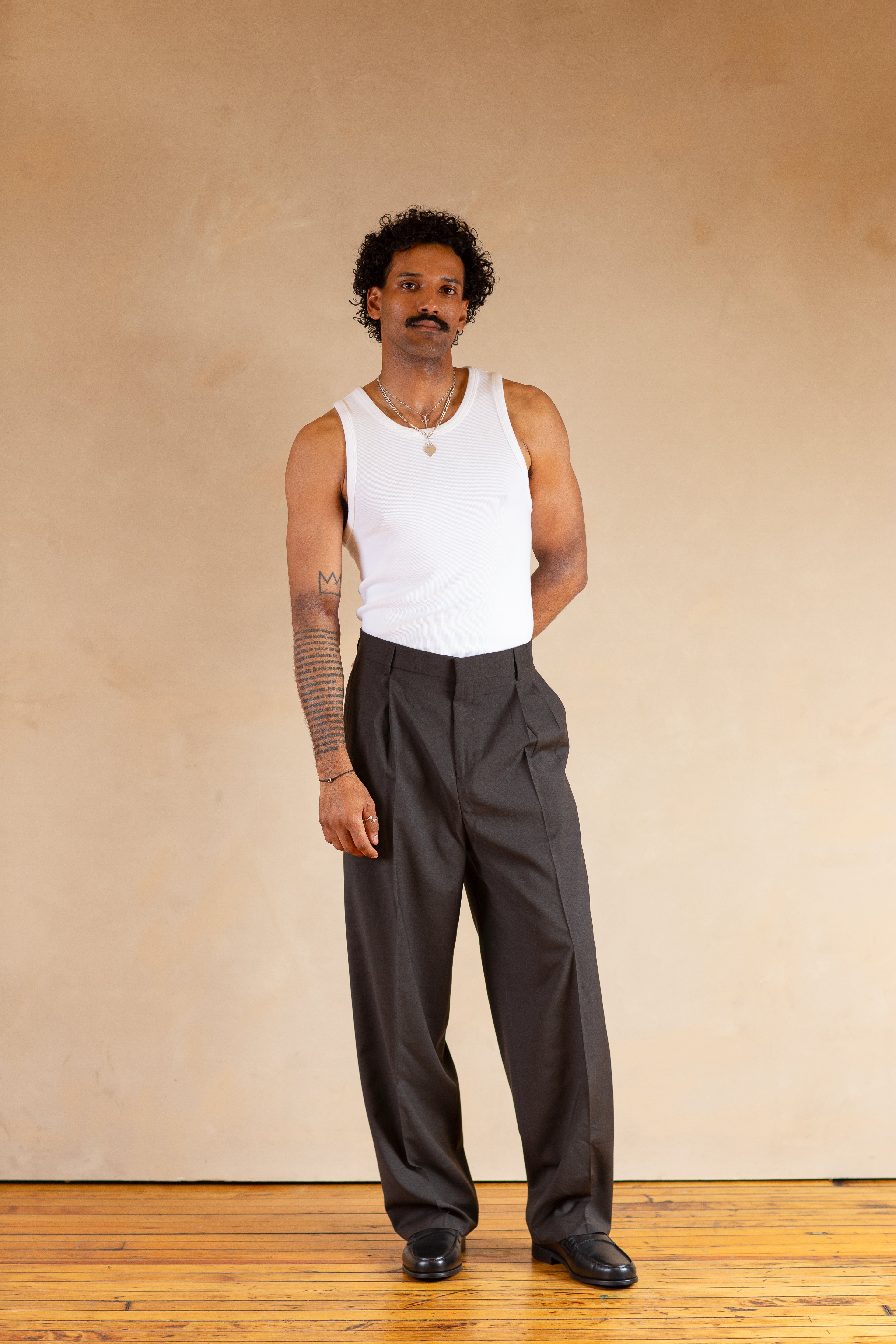 Man wearing a white tank top and Stone pants standing against a beige wall.